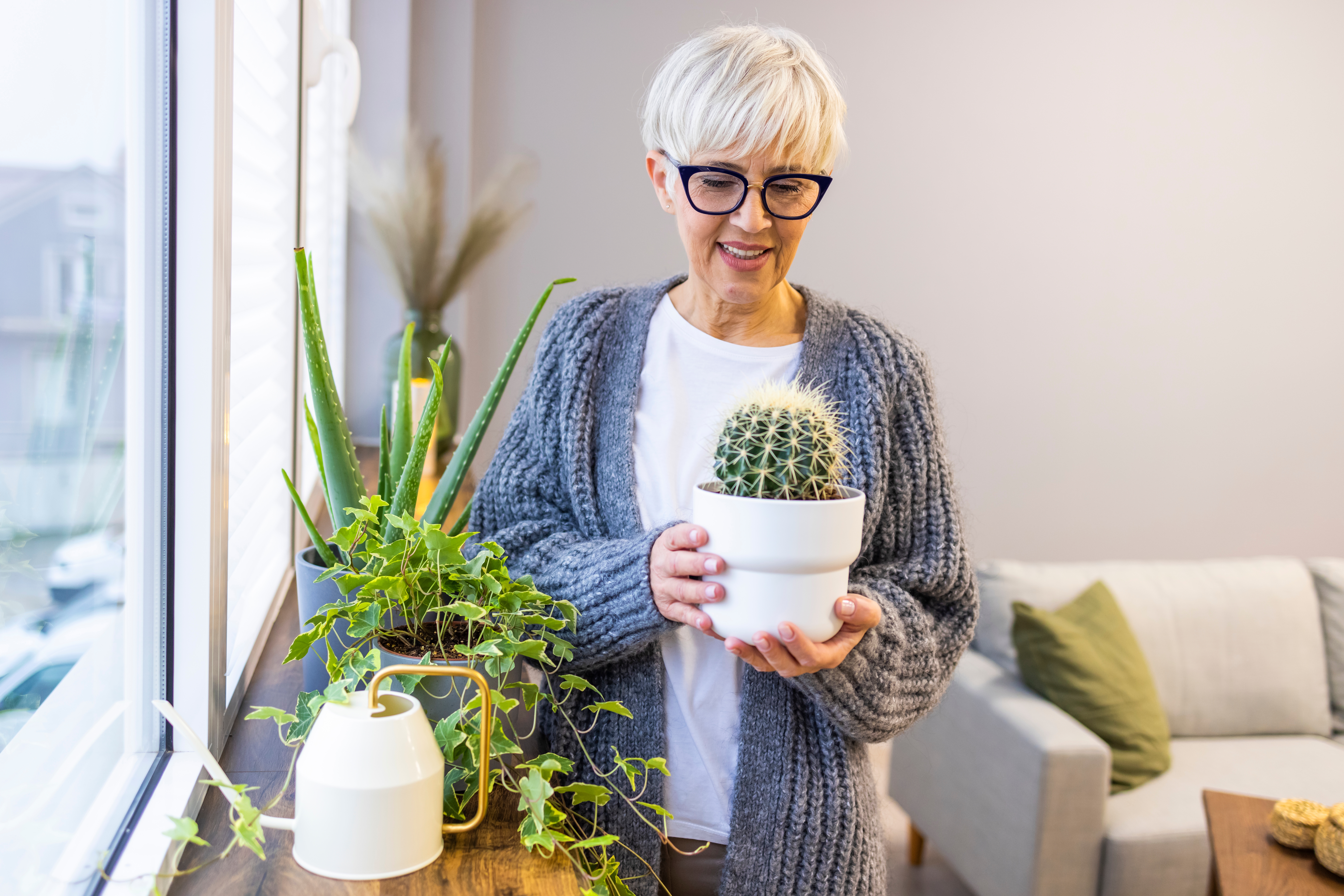 A senior woman holds a cactus near a shelf full of green house plants.